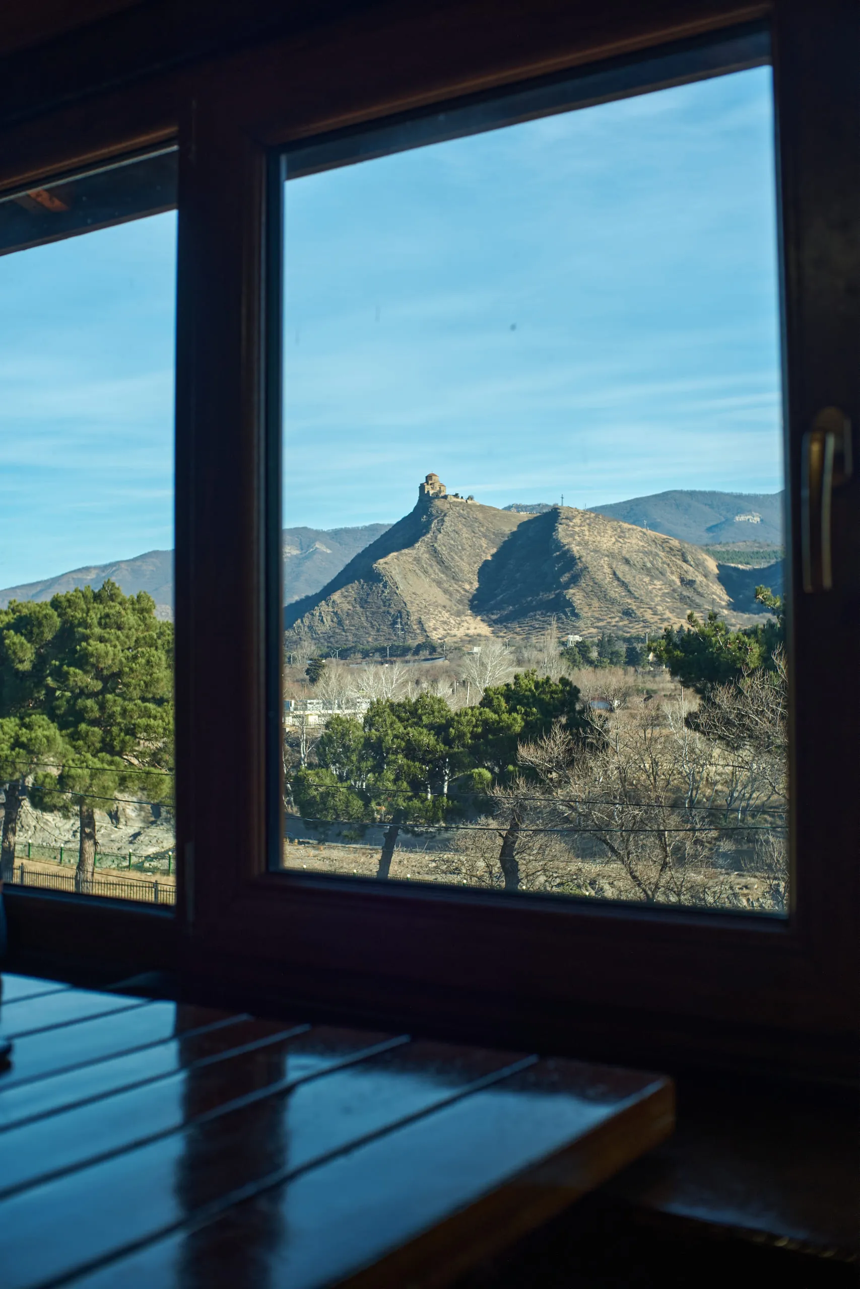 A large window in a wooden room framing a hill with a church on it
