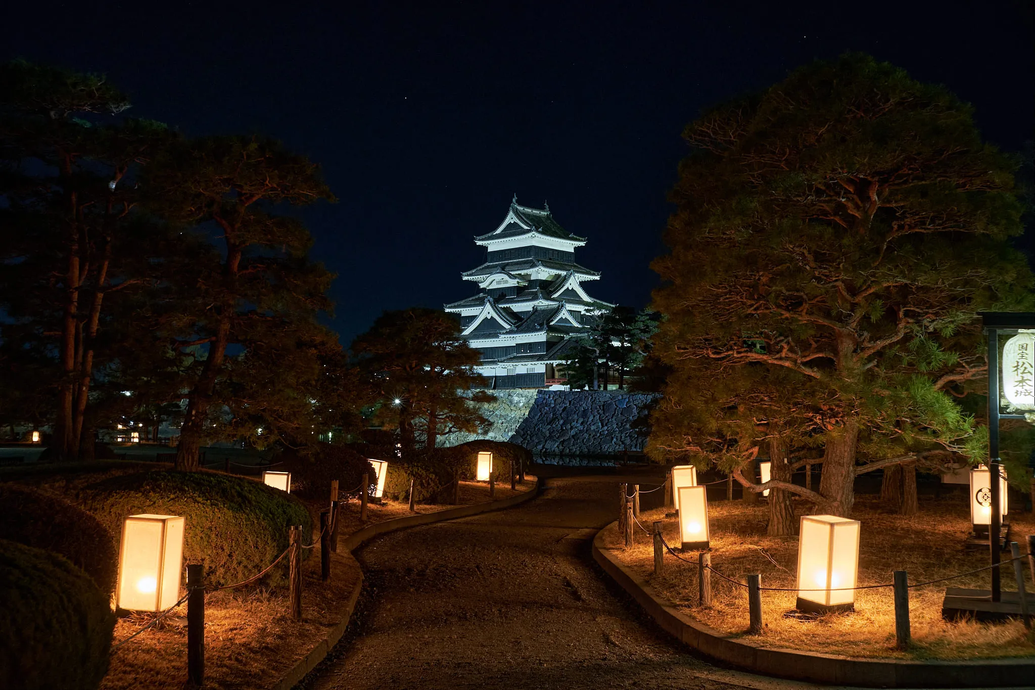 A castle keep floodlit at night behind pines, with paper lanterns lining the path in front of it