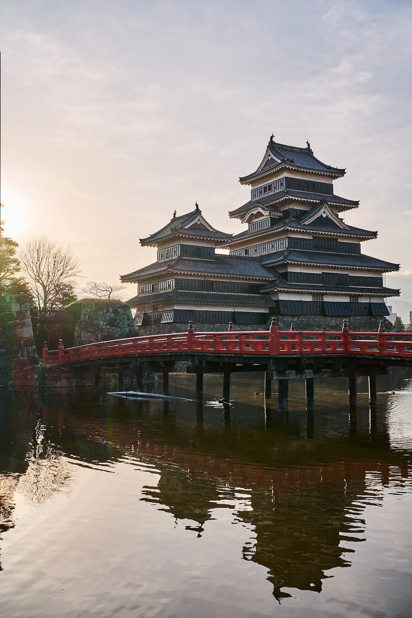 A black castle keep and a red arched bridge reflected in still water at sunrise