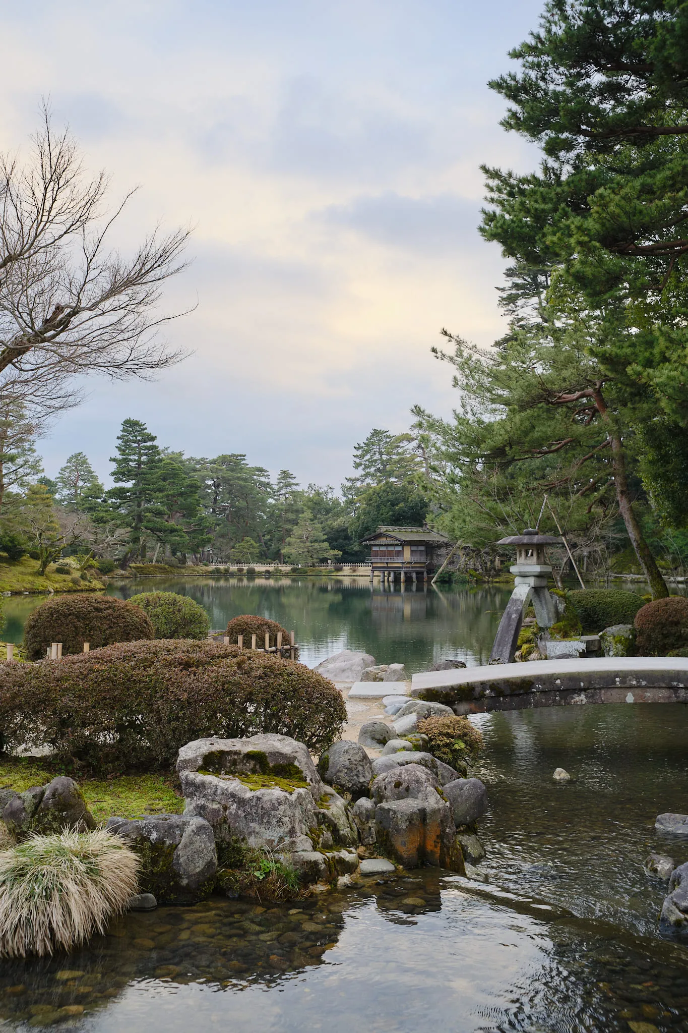A garden pond with clipped shrubs, a stone lantern and a low stone bridge, a wooden pavilion across the water