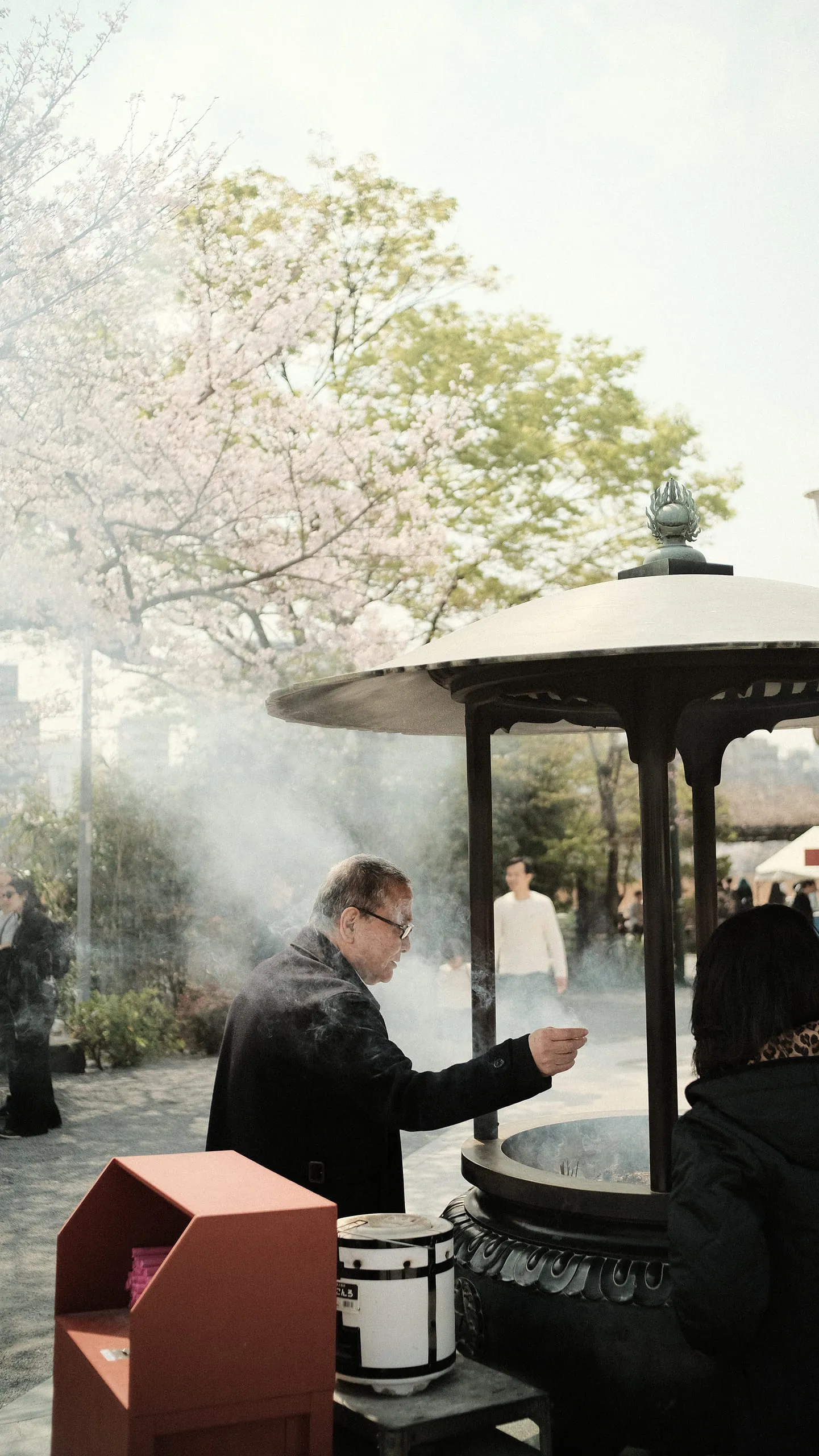 A man in a dark coat drawing incense smoke towards himself at a large bronze censer, cherry blossom behind