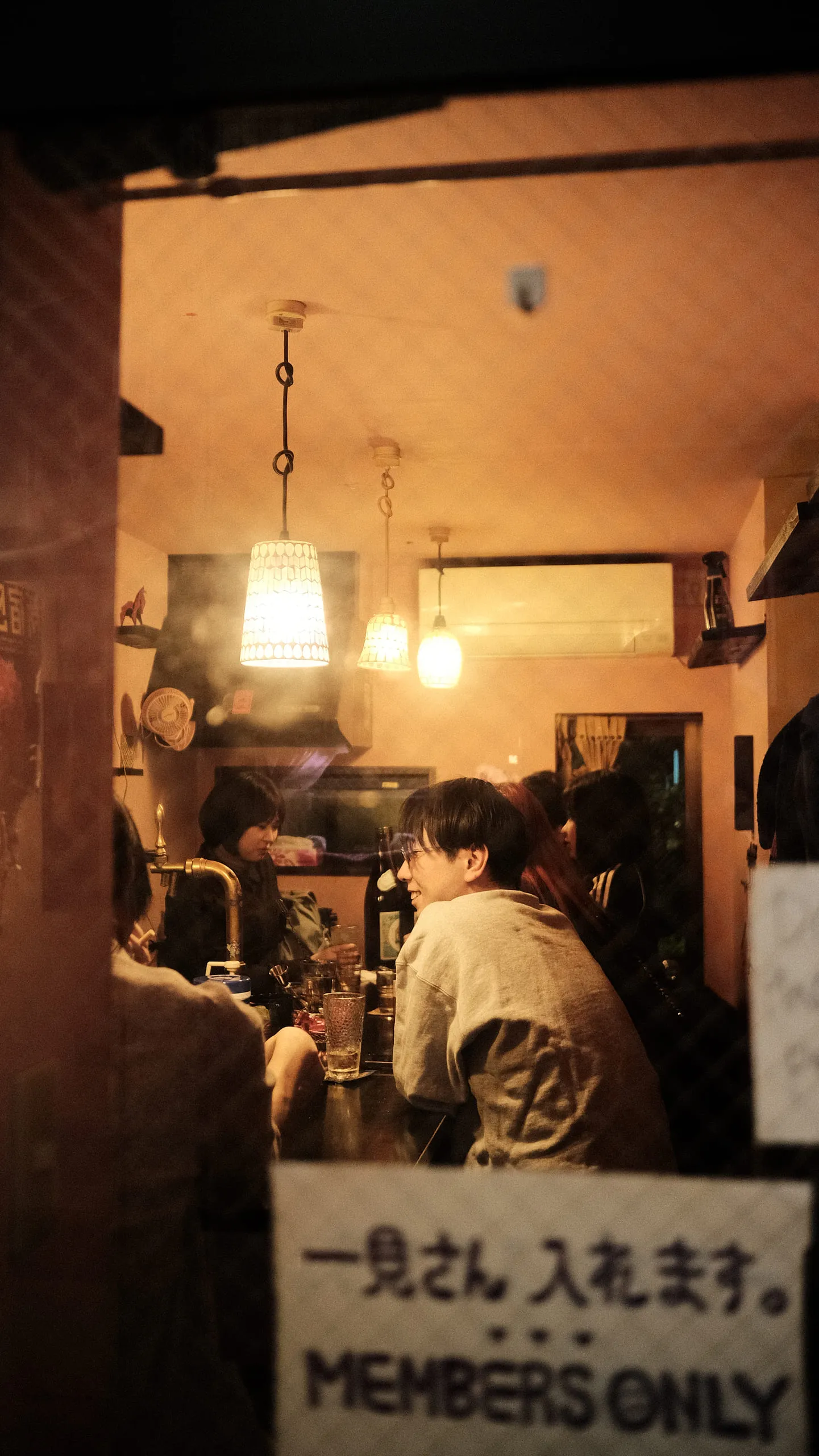 A small bar seen through a doorway, warm lamplight over a crowded counter, a handwritten sign in the foreground