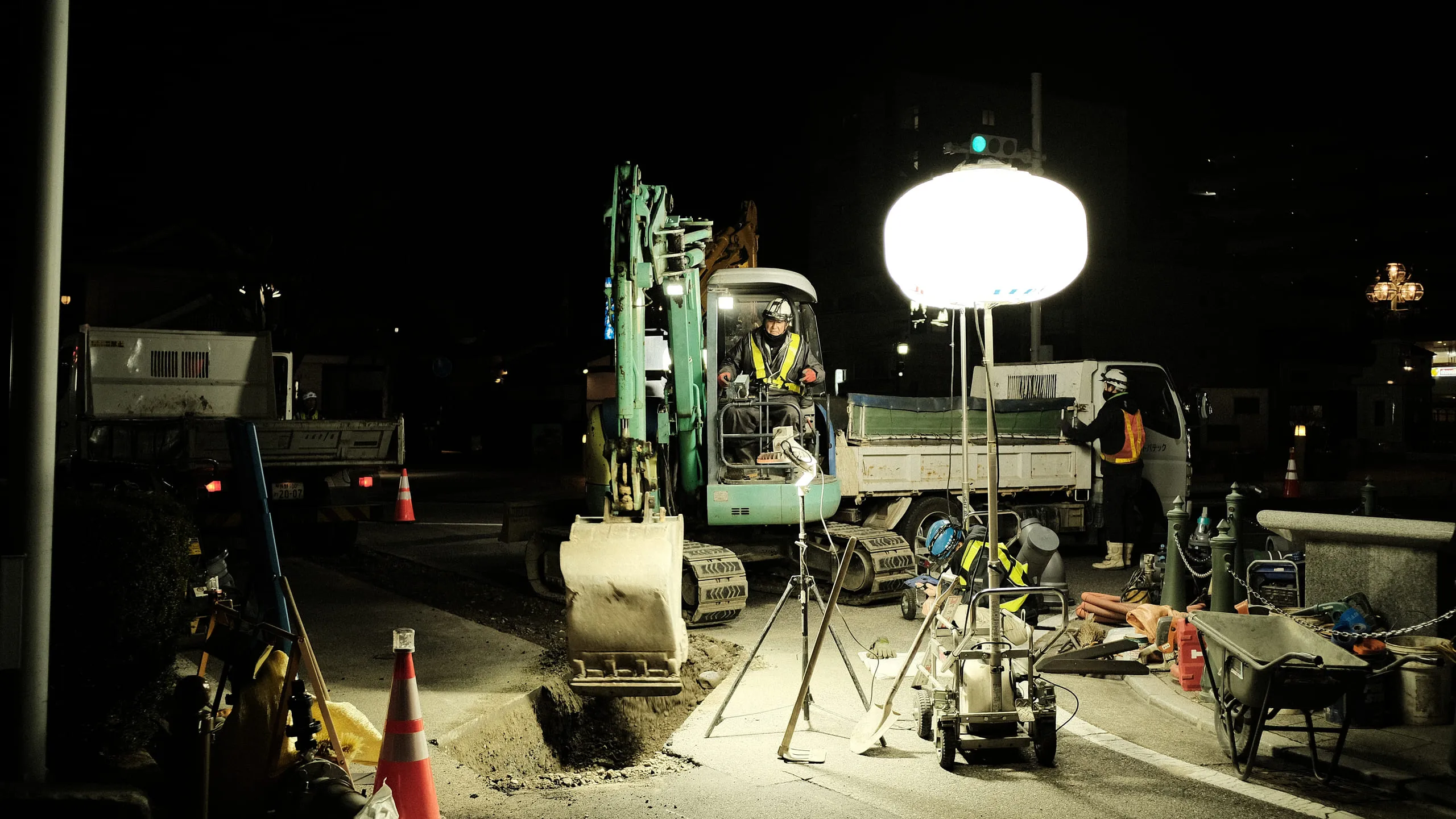 A road excavation at night lit by a large balloon lamp, with a digger, a tipper truck and workers in hi-vis
