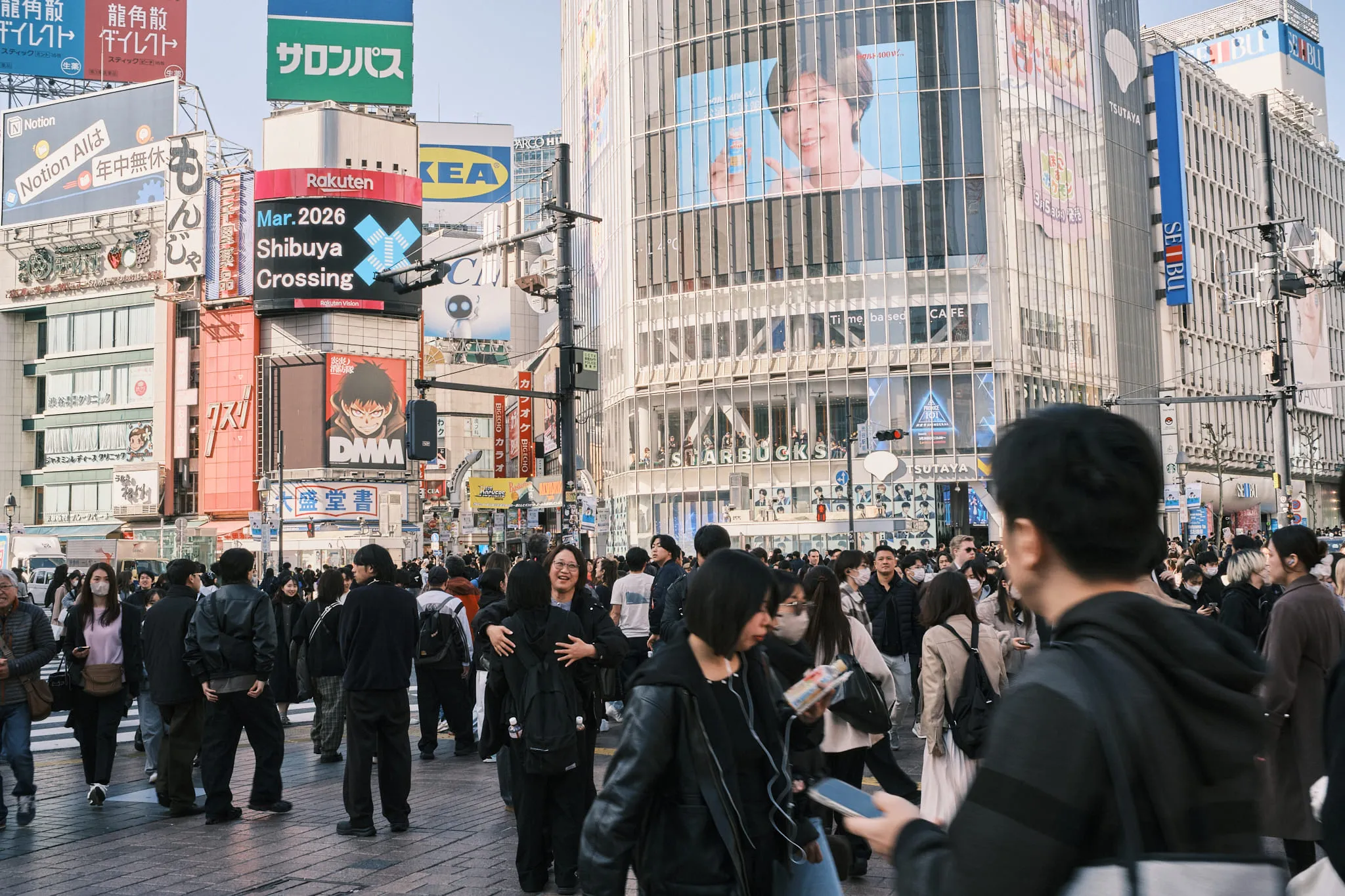 A dense crowd crossing a wide intersection below billboards and screens