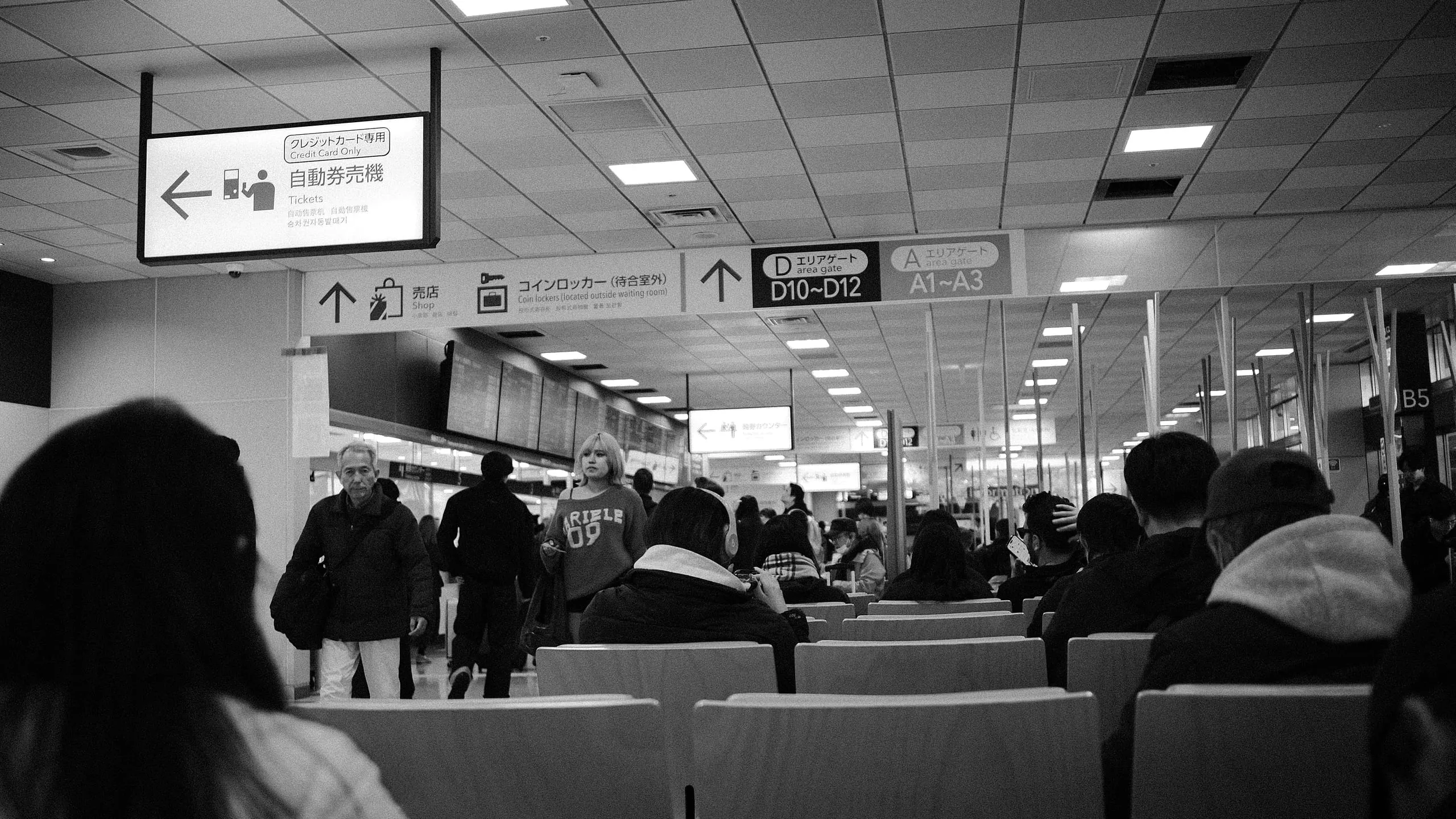 Black and white view of a station concourse, overhead signs pointing to ticket machines, shops, lockers and gates, with people waiting in rows of seats