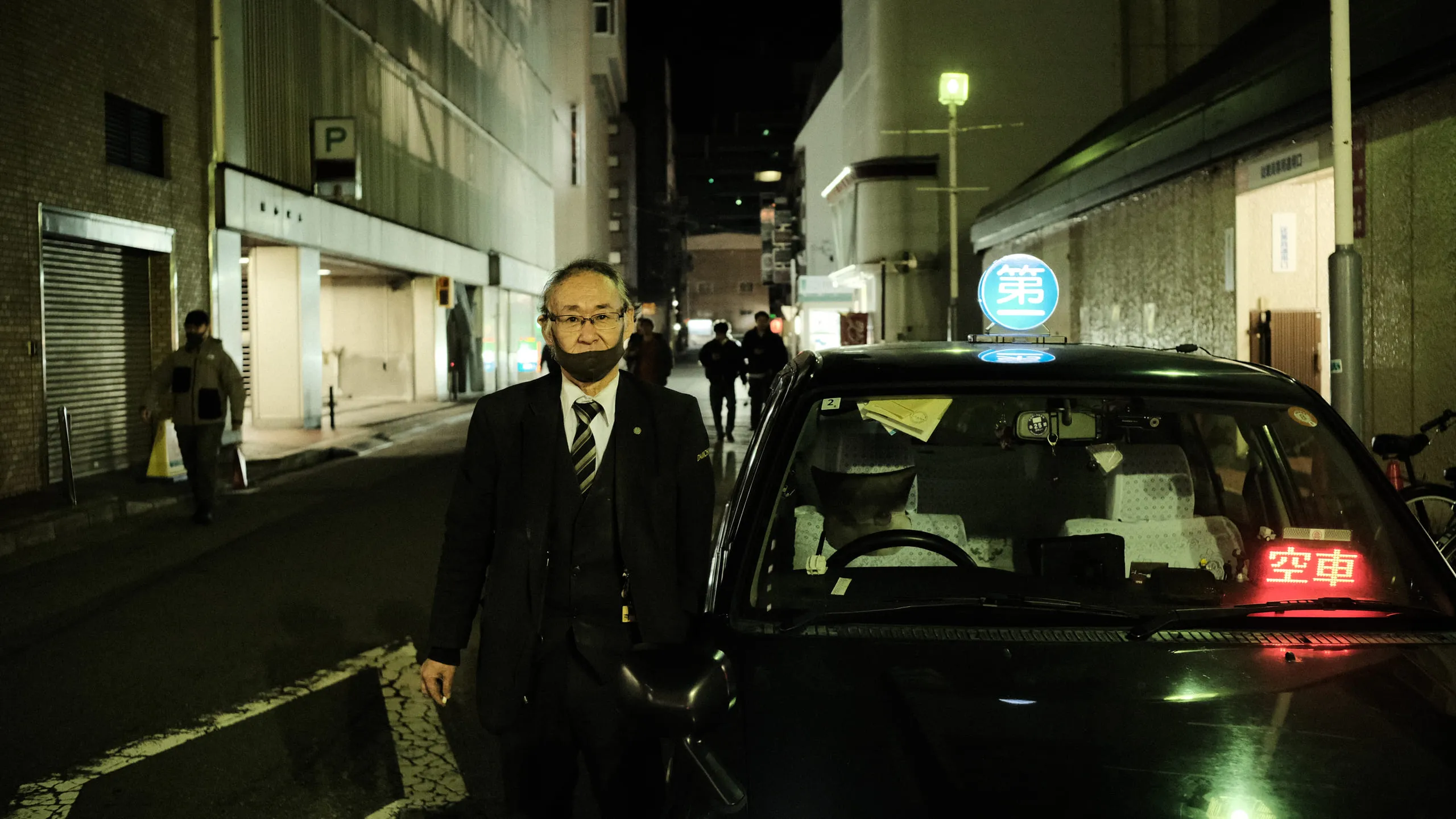 A taxi driver in a suit standing beside his cab on an empty street at night, the for-hire sign lit inside the windscreen