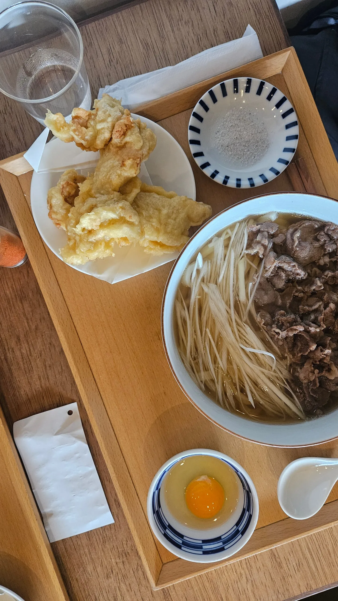 A wooden tray holding a bowl of noodle soup with beef and enoki, a plate of tempura, and a raw egg in a small bowl